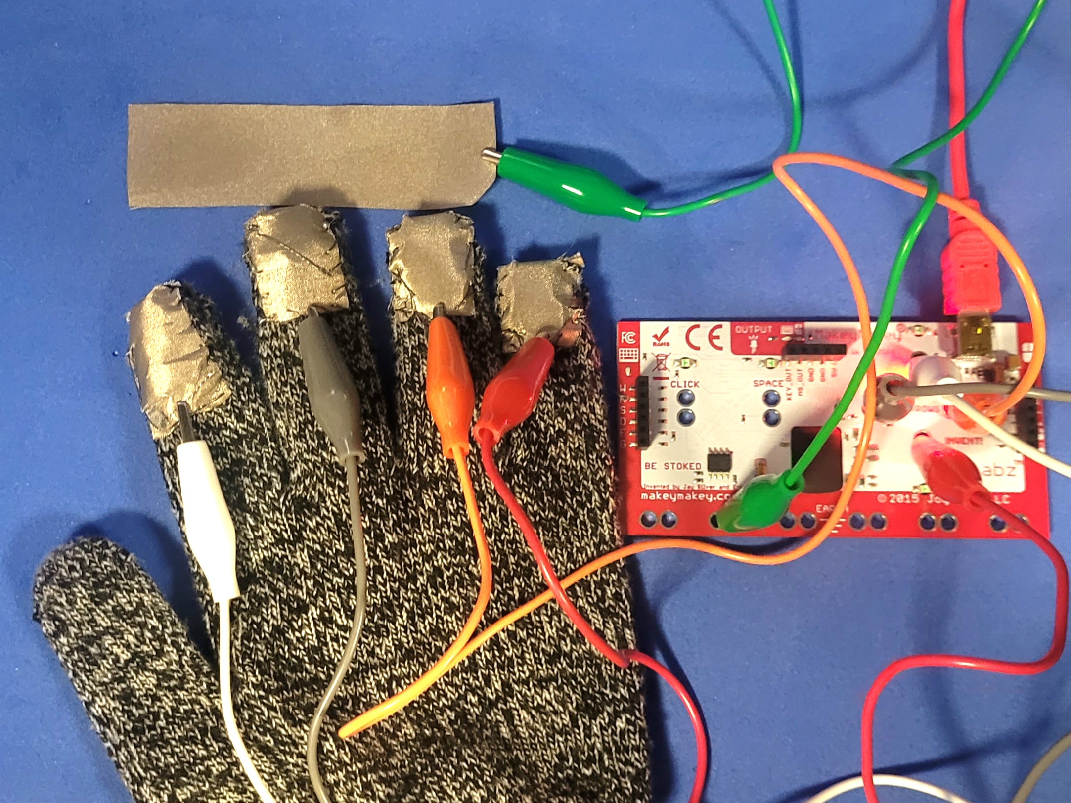 A photo of circuit that uses a conductive glove made for project 2 but with the inputs reversed, each finger corrosponds to a key input on the MakeyMakey while the conductive fabric acts as the ground.